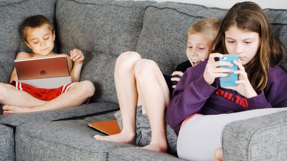 Drei Kinder sitzend auf einem Sofa, schauen auf Handy und iPad