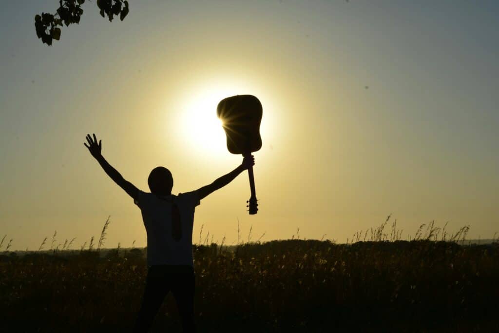 Mensch steht auf einem Feld draußen, mit Hände gespreizt nach oben, an einer Hand hält die Person eine Gitarre, sonniger Himmel