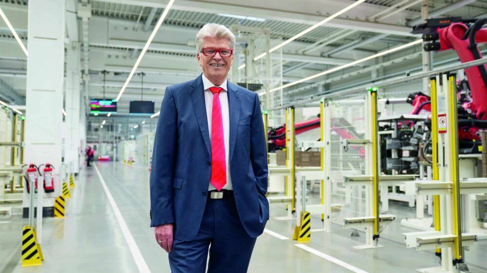 Friedhelm Loh steht in einem Produktionswerk. Er trägt eine Krawatte und einen Anzug.
