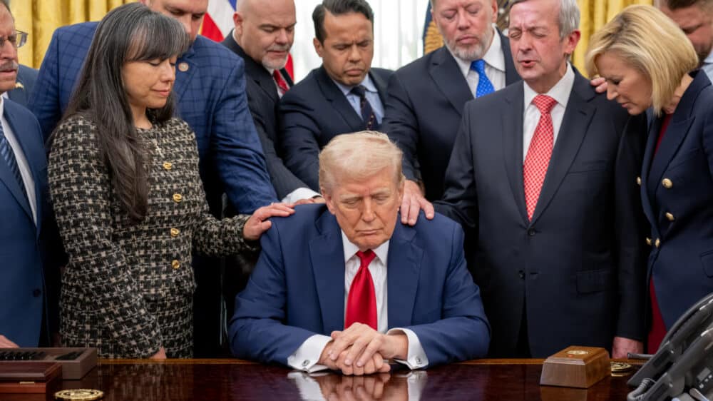 Donald Trump sitzt am Schreibtisch im Oval Office. Geistliche legen ihm die Hände auf und beten für ihn.