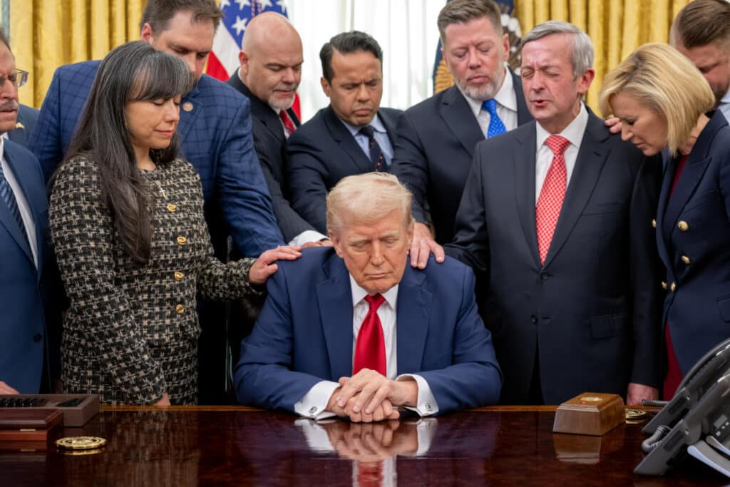 Donald Trump sitzt am Schreibtisch im Oval Office. Geistliche legen ihm die Hände auf und beten für ihn.