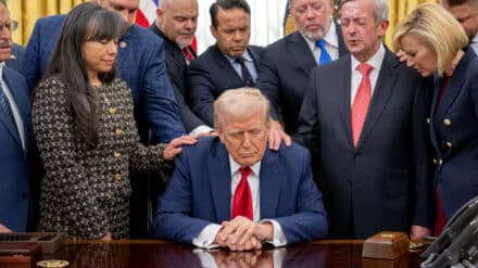 Donald Trump sitzt am Schreibtisch im Oval Office. Geistliche legen ihm die Hände auf und beten für ihn.