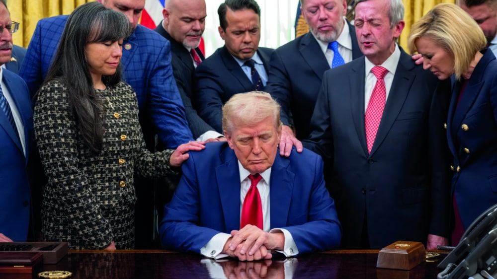 Donald Trump sitzt am Schreibtisch im Oval Office. Geistliche legen ihm die Hände auf und beten für ihn.