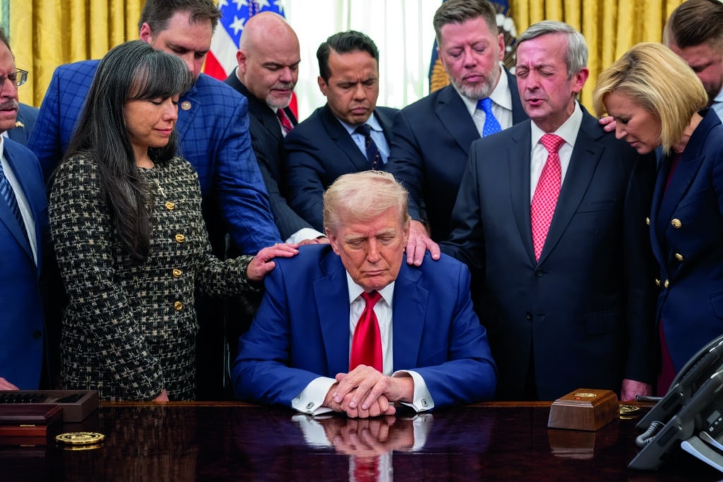 Donald Trump sitzt am Schreibtisch im Oval Office. Geistliche legen ihm die Hände auf und beten für ihn.